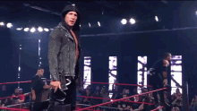 a wrestler in a hooded jacket stands in a wrestling ring pointing at the camera