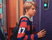 a young boy wearing a striped sweater with the number 87 on the back is standing in front of a purple locker .