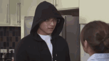 a man wearing a hooded jacket and glasses is talking to a woman in a kitchen .
