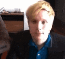 a man with blonde hair is wearing a blue shirt and a black jacket and looking at the camera .