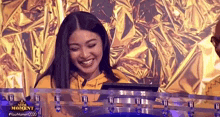 a woman in a yellow hoodie is smiling while sitting in front of a gold foil wall .