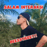 a man wearing sunglasses and a baseball cap stands in front of palm trees with the words salam interaksi beesstieeee !!! below him