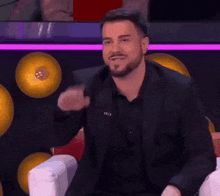 a man in a suit and black shirt is sitting in a chair with his hand up .