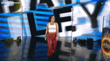 a woman in a white tank top and red pants is walking on a stage in front of a sign that says ley