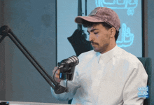 a man wearing a hat and a white shirt is sitting in front of a microphone with arabic writing on the bottom