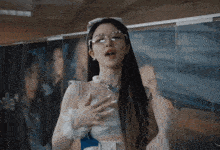 a woman wearing glasses and a pearl necklace stands in front of a wall with a picture of a woman behind her