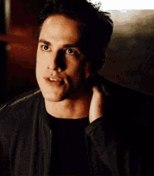 a man in a black jacket is holding his hand to his ear and looking at the camera .