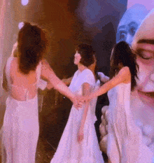 a group of women in white dresses are dancing in front of a painting of a man 's face .