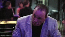 a man in a suit and black shirt is sitting at a table in a dark room .