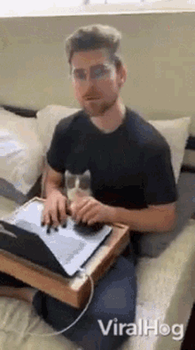 a man is sitting on a bed using a laptop and a cat is sitting on a tray next to him .