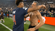 a shirtless man is hugging another shirtless man on a soccer field with a canal + sign in the background