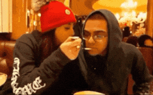 a woman is feeding a man a spoon while wearing a black sweatshirt with the word chrome on the sleeves