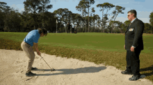 a man in a suit watches a man in a blue shirt putt a golf ball from a bunker