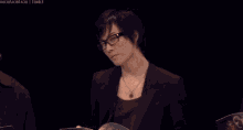 a man wearing glasses and a necklace is sitting in front of a microphone and talking into it .