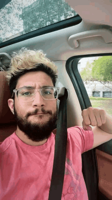 a man with a beard wearing glasses and a pink shirt is sitting in a car