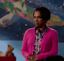 a woman wearing a pink cardigan and a floral shirt is standing in front of a painting .