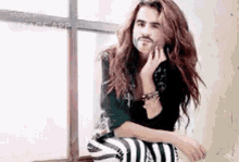 a man with long hair is sitting in front of a window .