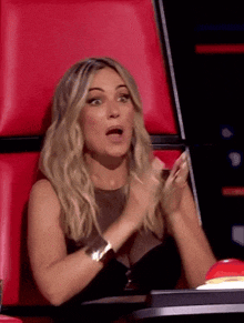 a woman in a black dress is sitting in a red chair and clapping her hands