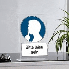 a sign with a silhouette of a man holding his finger to his mouth is on a table next to a plant .