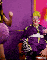 a drag queen in a purple outfit is sitting in a chair