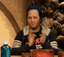 a man with blue hair is sitting at a table with his hands folded in front of him .