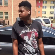 a young man wearing a black metallica t-shirt stands in front of a car .