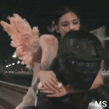 a woman wearing angel wings kisses a man wearing a helmet with the letters ms on the bottom right