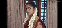 a woman in a red saree is standing in front of a window wearing a garland of flowers .