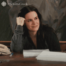 a woman sits at a desk with her hand on her forehead and the word schitts creek is on the bottom