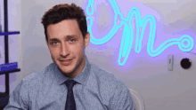 a man in a blue shirt and tie is sitting in front of a neon sign that says on