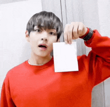 a young man in a red sweater is holding a piece of paper in front of his face .