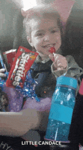 a little girl is sitting in a car seat holding a lollipop and a bag of skittles