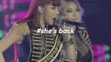 two women are singing into microphones on a stage and the words `` she 's back '' are on the screen .