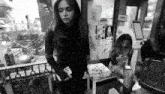 a black and white photo of a woman standing next to a woman sitting on a chair .