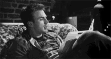 a man is laying on a couch reading a book and smoking a cigarette