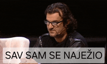 a man wearing glasses and a leather jacket is sitting in front of a sign that says sav sam se najezio