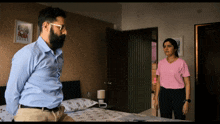 a man with a beard stands next to a woman in a pink shirt in a bedroom