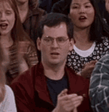 a man wearing glasses is sitting in a crowd of people clapping .