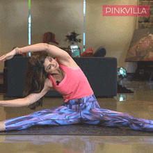 a woman in a pink tank top and purple pants is stretching on a yoga mat in front of a sign that says pinkvilla