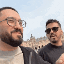 two men with glasses and a beard are standing next to each other in front of a building