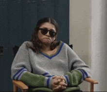 a woman wearing a sweater and sunglasses is sitting in a chair with her hands folded .