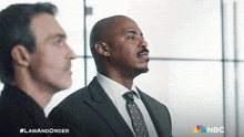 a man in a suit and tie stands next to another man in a suit and tie with a nbc logo behind them