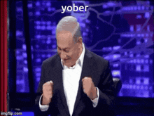 a man in a suit is sitting on a stage with his fist in the air and the words yober written on the bottom .
