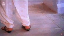 a woman wearing white pants and black shoes is walking on a wooden floor .