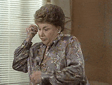 an older woman in a floral shirt is standing in front of a window