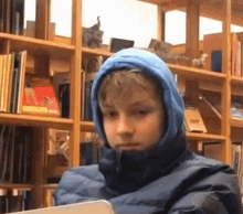 a young boy wearing a blue hooded jacket is sitting in front of a bookshelf in a library .