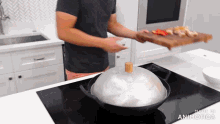 a man in a black shirt is holding a cutting board and a wok with a wooden lid on it