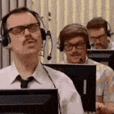 a man with a mustache is wearing headphones and glasses while sitting at a desk in front of a computer .