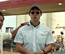 a man wearing sunglasses and a storm chasers shirt is being helped by another man
