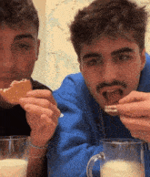 two men are sitting at a table eating cookies and drinking milk .